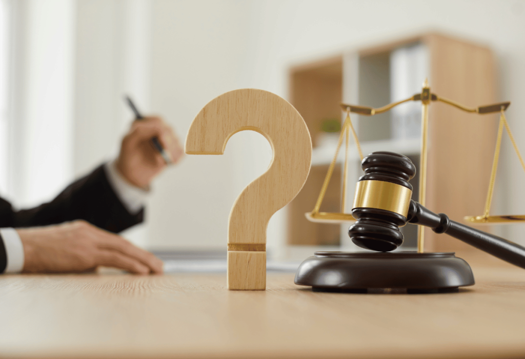 Martelo de juiz e balança da justiça sobre uma mesa, ao lado de um grande ponto de interrogação de madeira, simbolizando falhas ou erros no processo jurídico.