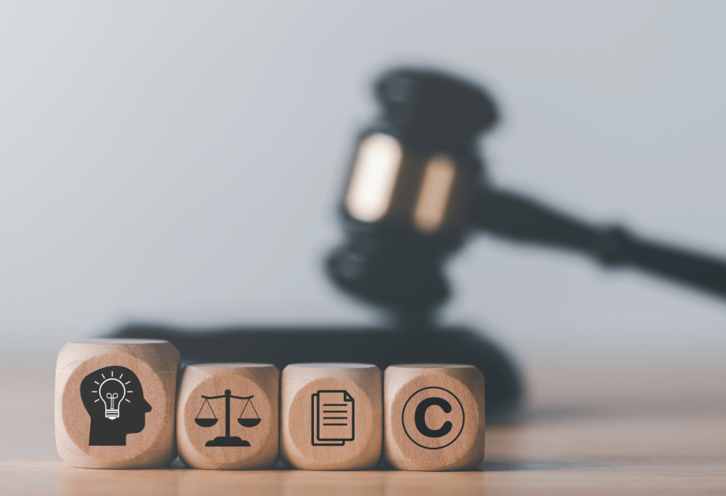 Blocos de madeira com ícones de ideia, balança da justiça, documentos e copyright diante do martelo de juiz ao fundo, representando propriedade intelectual e proteção jurídica.